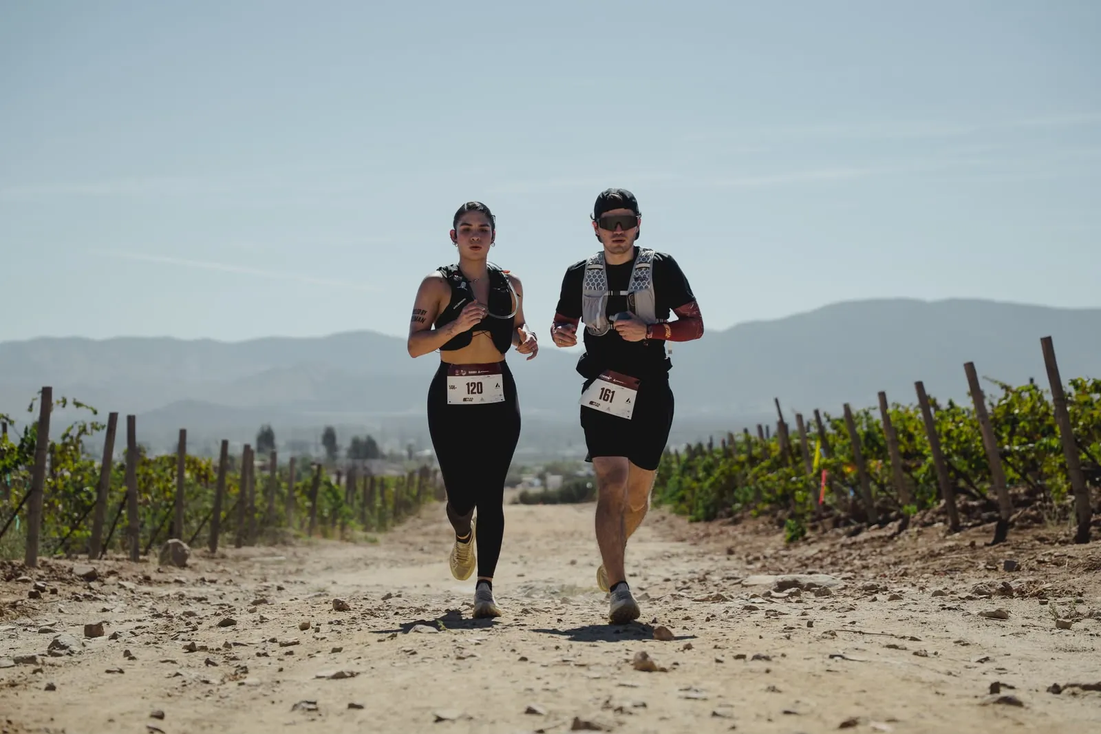 Two runners with race bibs approaching on a vineyard road.