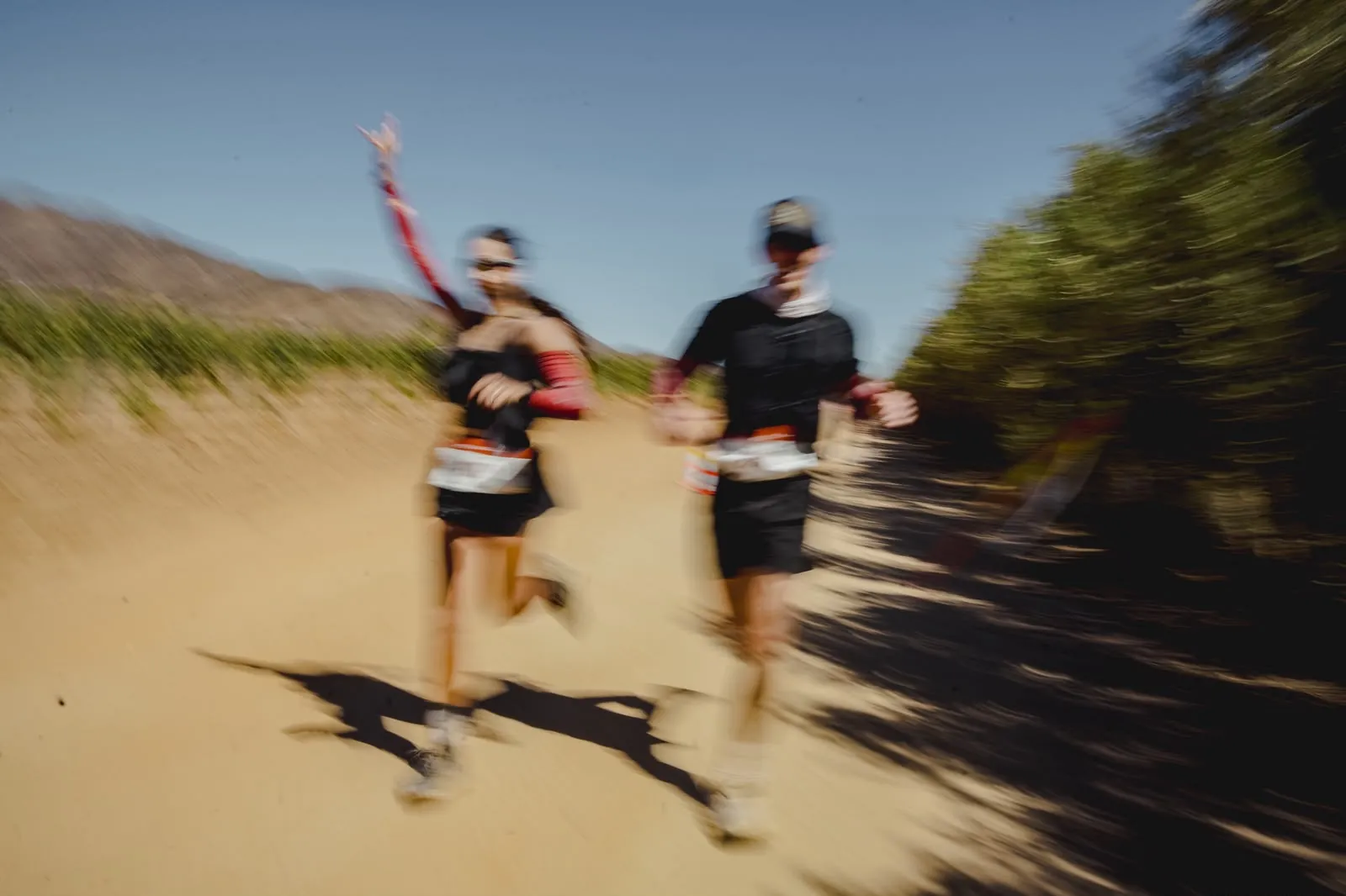Two runners waving mid-race, motion-blurred, vineyard behind.