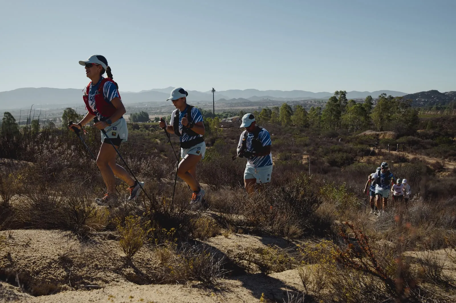 A line of runners with trekking poles, hiking a ridge above the valley.